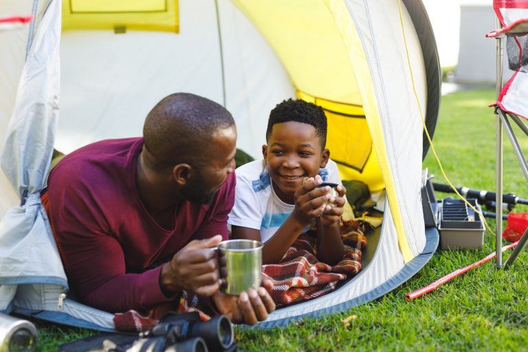 African american father with son having fun and lying in tent with drinks in garden