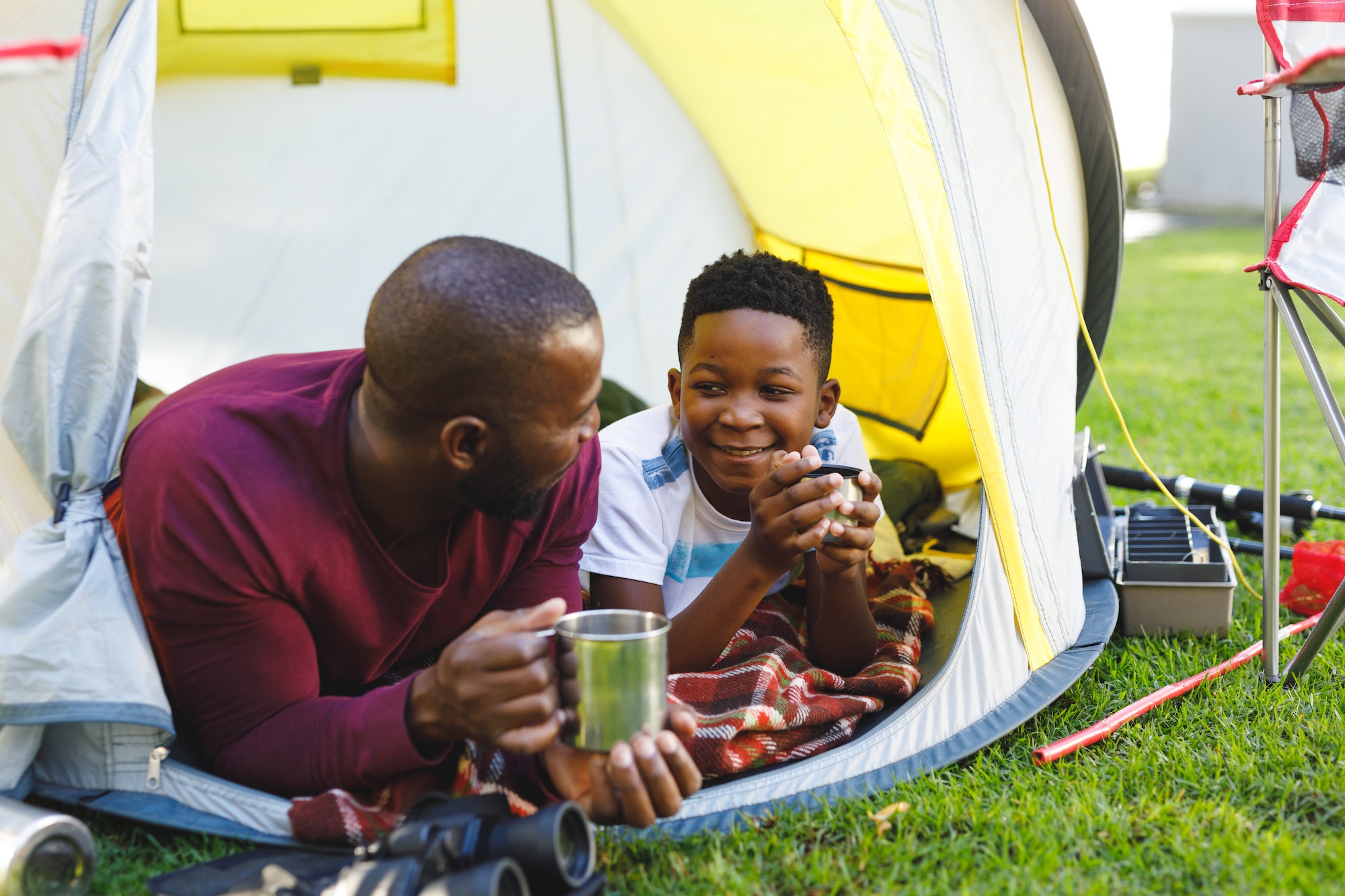 African american father with son having fun and lying in tent with drinks in garden